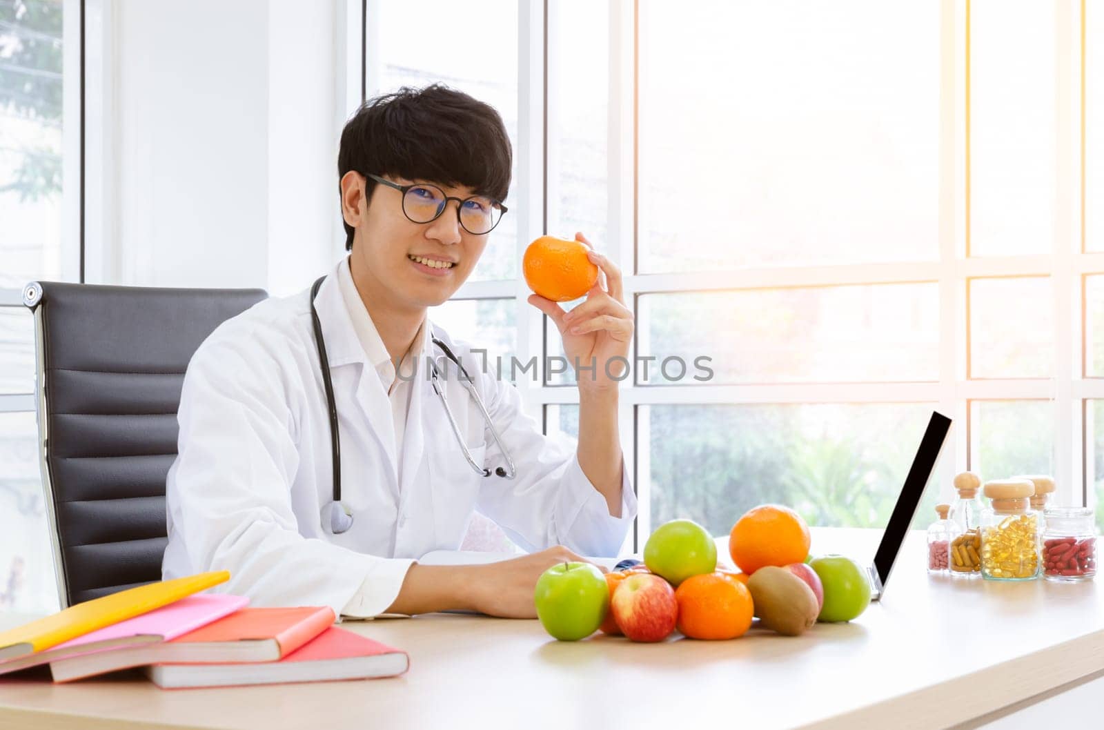 Male nutritionist in white lab coat holding orange fruit in clinic room with fresh variaty fruits and vitamins on his desk. Healthy eating with fruit and vitamin C concept to prevent the flu.