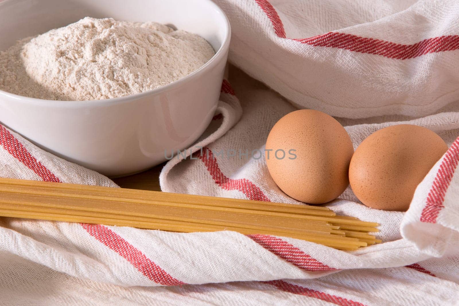 Gourmet close up with a bowl full of flour, spaghetti, and two eggs in white kitchen cloths with red border, on a wooden board-