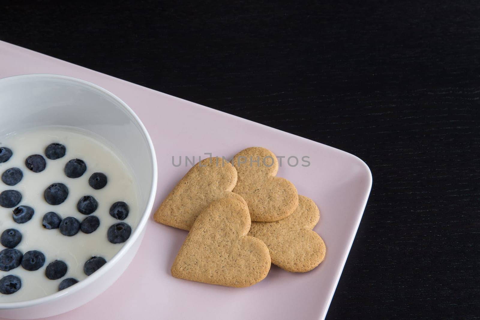 Breakfast. Yogurt with blueberries heart shaped cookies. Ingredients in a pink tray on a black wood table. Detail from above with copy space.