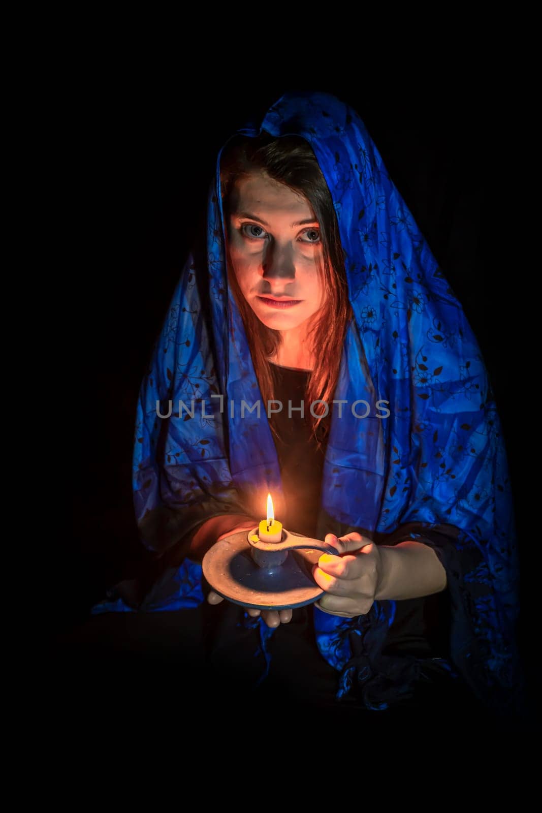 Sad young woman with candle and blue headscarf against black background