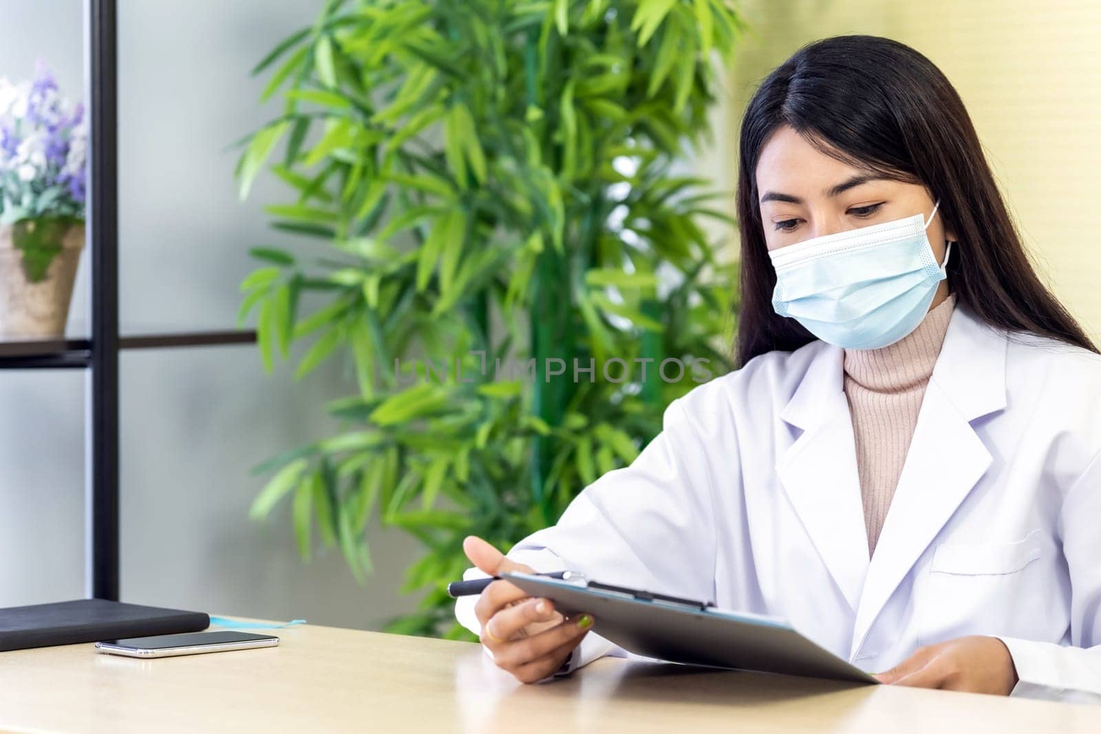 Portrait of asian female doctor wear protective face mask sit in her office room in hospital clinic and reading on patience file before checking patience. Health care Concept.