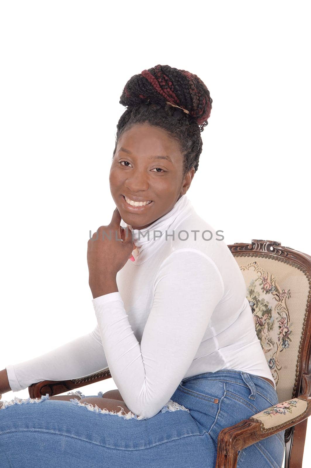A smiling African woman sitting in a white top and jeans in an.old armchair, looking into the camera, isolated for white background.