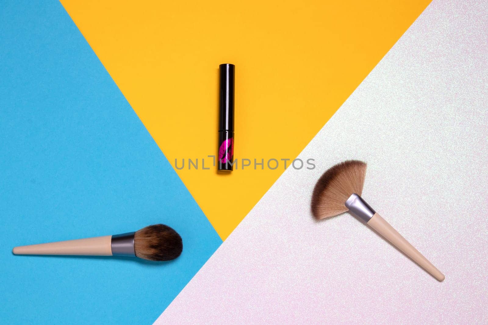 Two professional makeup brushes and a lipstick flat lay on a bright pink, yellow and blue background with copy space. Template for cosmetics product display montage or advertising text. Beauty and skincare concept. Top view.