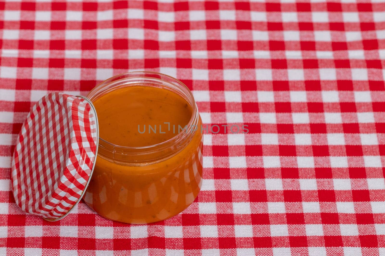 Closeup of a jar with home made classical sauce of fresh paprika with tomatoes, horseradish, garlic and basil on a red checkered tablecloth. Template for food product display montage or advertising text. Concept of healthy nutrition. Macro.
