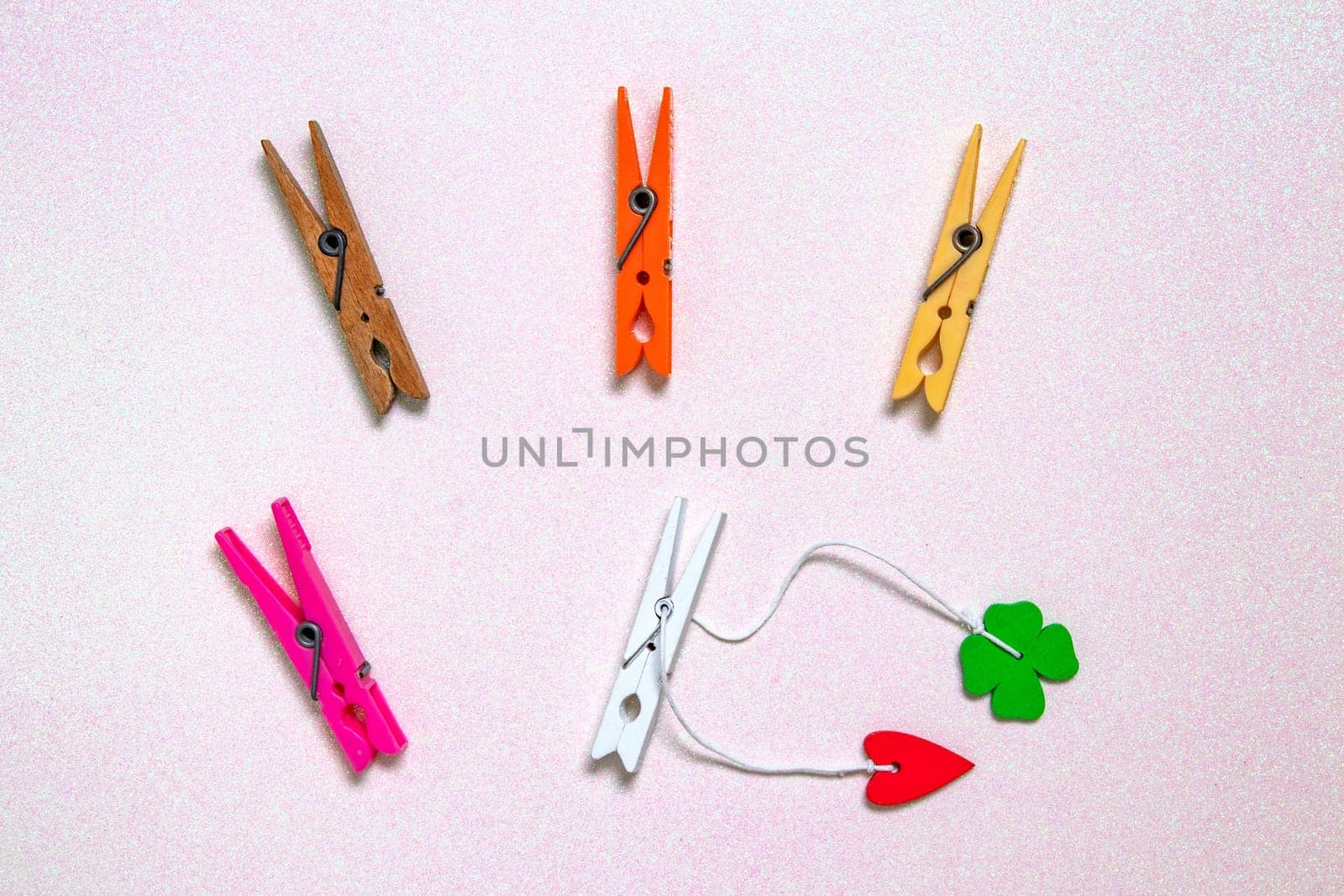 Colorful set of five wooden and plastic clothespins on a light pink glitter sparkle background with space. A red heart and a green lucky clover leaf are attached to the white wooden clip. Topview.