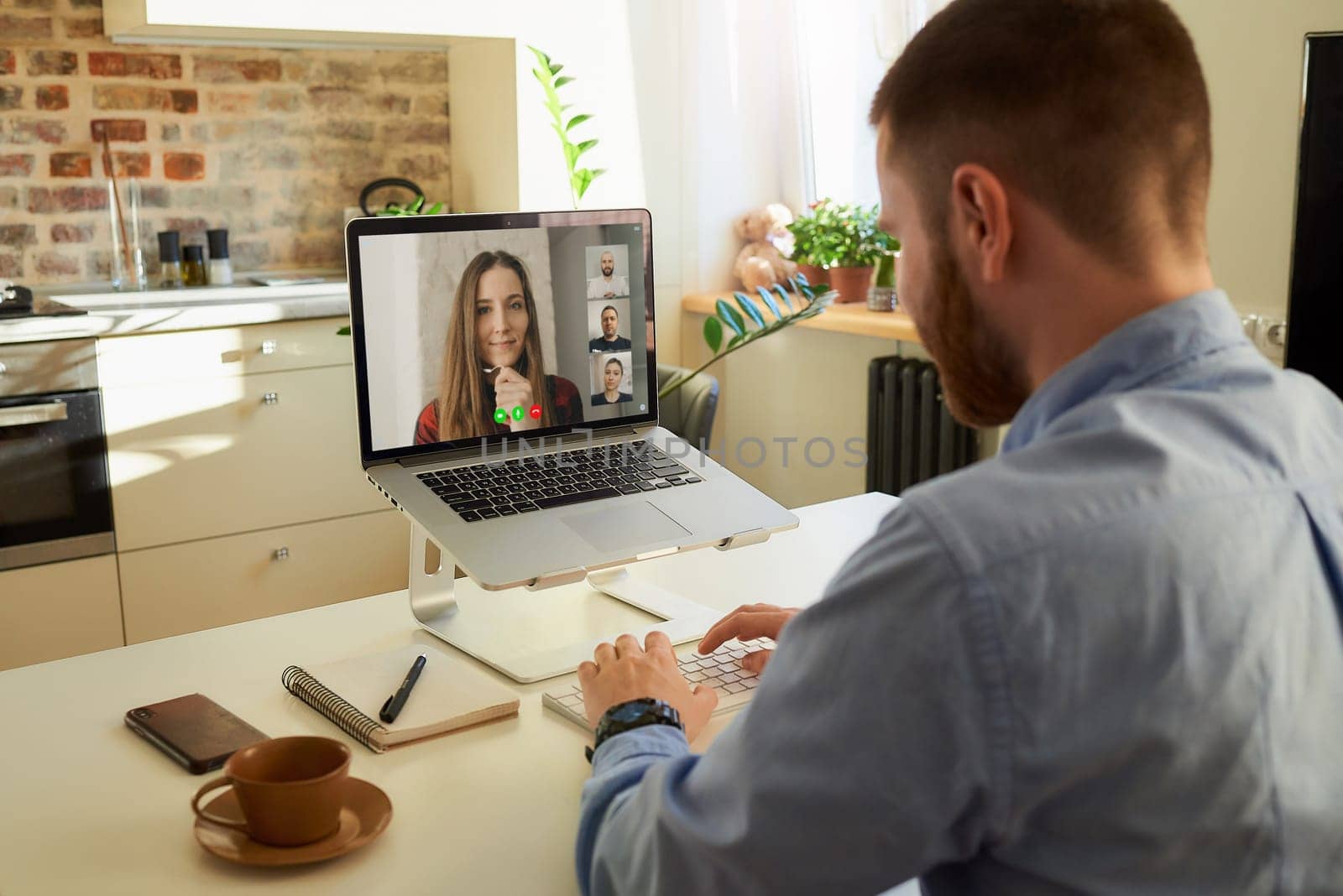 Back view of a male employee who works remotely talking to his colleagues about a business by a video conference on a laptop computer at home. A multiethnic business team at an online meeting.