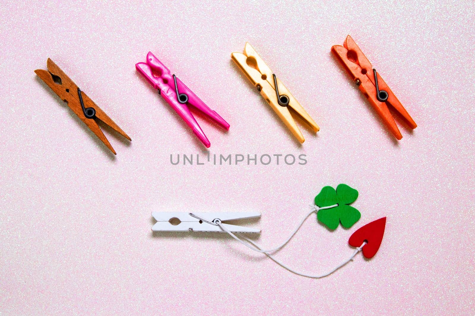 Colorful set of five wooden and plastic clothespins on a light pink glitter sparkle background with space. A red heart and a green lucky clover leaf are attached to the white wooden clip. Topview.