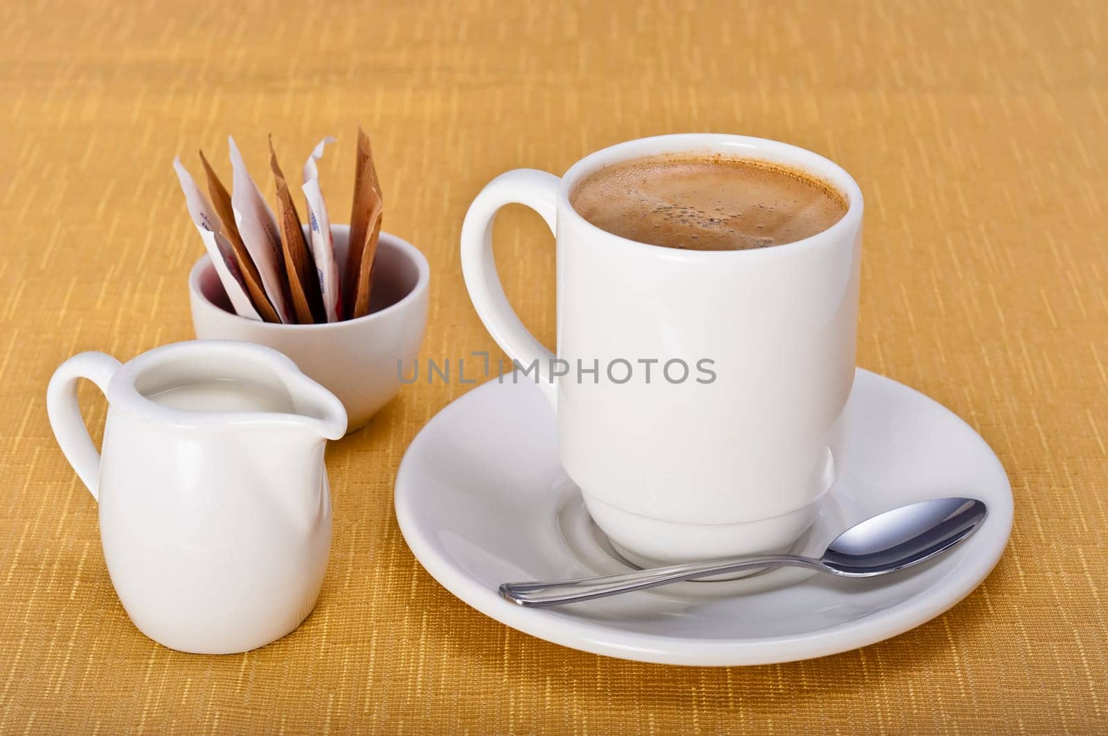 Mug of coffee with saucer, teaspoon, milk jug and sugar sachets on a golden table cloth