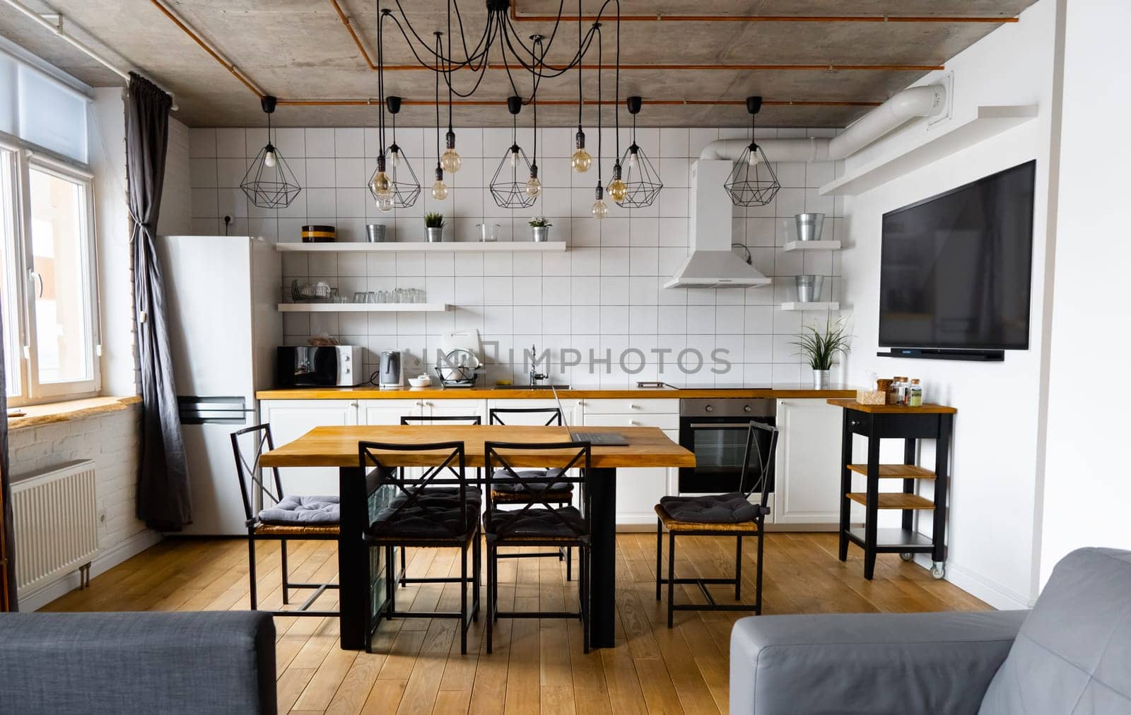 An eat-in kitchen interior design in modern scandinavian style with big wooden table and chairs against light wood floor, bright white walls and furnitures with TV, appliances and hanging light bulbs