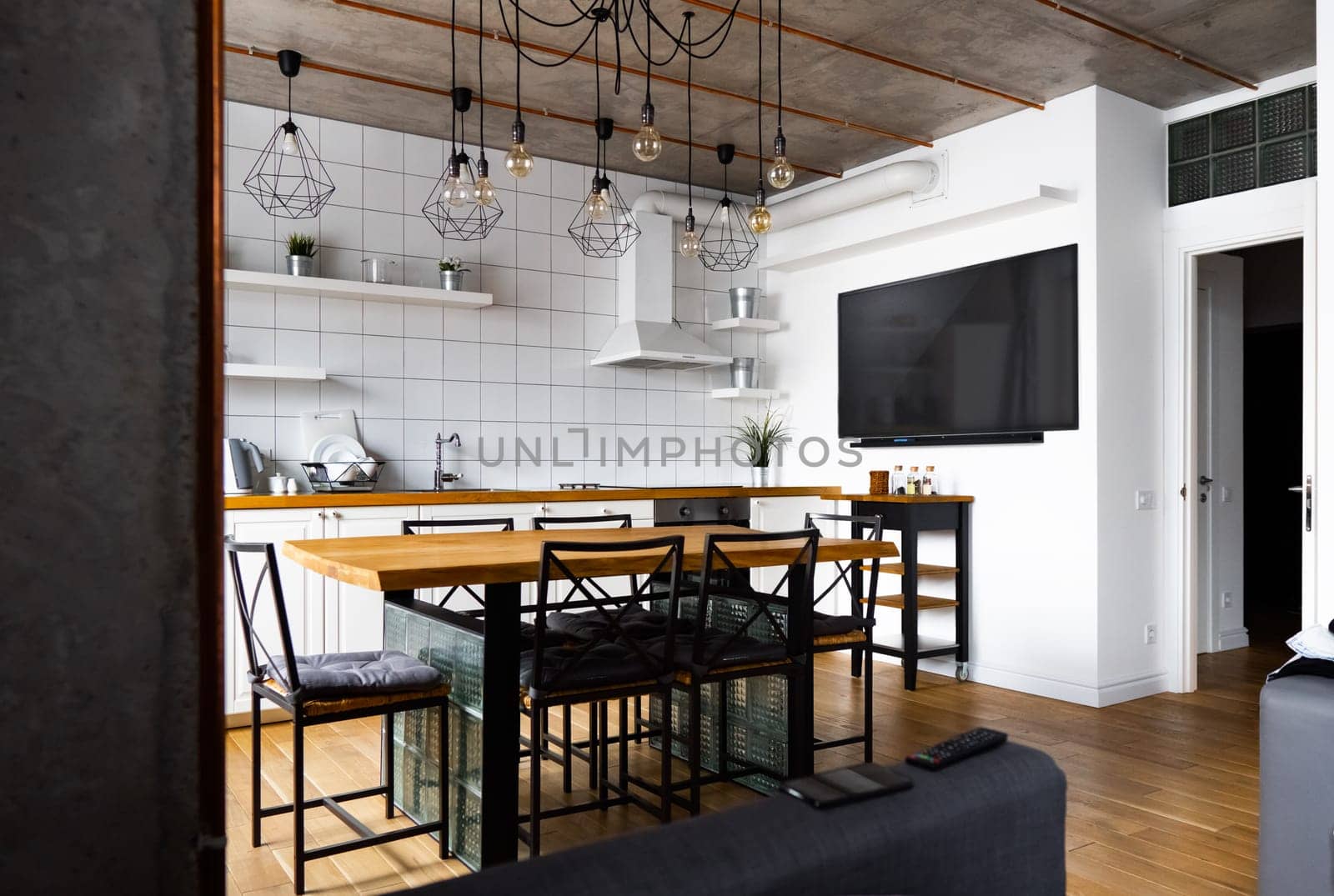 An eat-in kitchen interior design in modern scandinavian style with big wooden table and chairs against light wood floor, bright white walls and furnitures with TV, appliances and hanging light bulbs