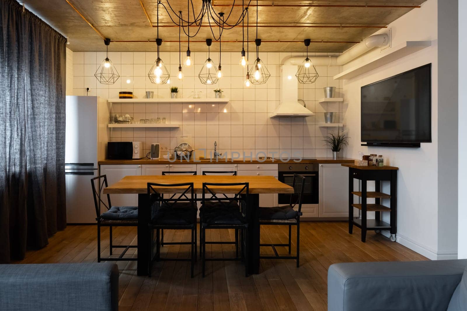 An eat-in kitchen interior design in modern scandinavian style with big wooden table and chairs against light wood floor, bright white walls and furnitures with TV, appliances and hanging light bulbs