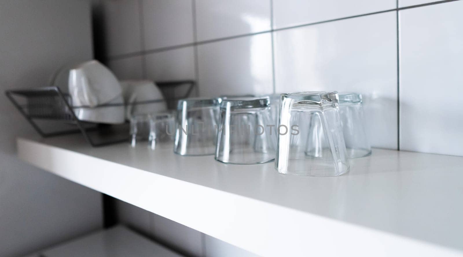 White wooden shelf and budget lightweight dish rack with dishes, cups and glasses against wall tiles at modern scandinavian kitchen. Wide kitchen shelves and wall shelving for storage and display