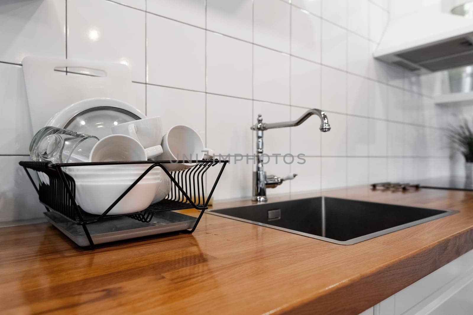 Budget and lightweight antimicrobial dish drainer with drain board at modern scandinavian kitchen. Dish rack holds many dishes and cups against wooden countertop, white wall tiles, sink and faucet
