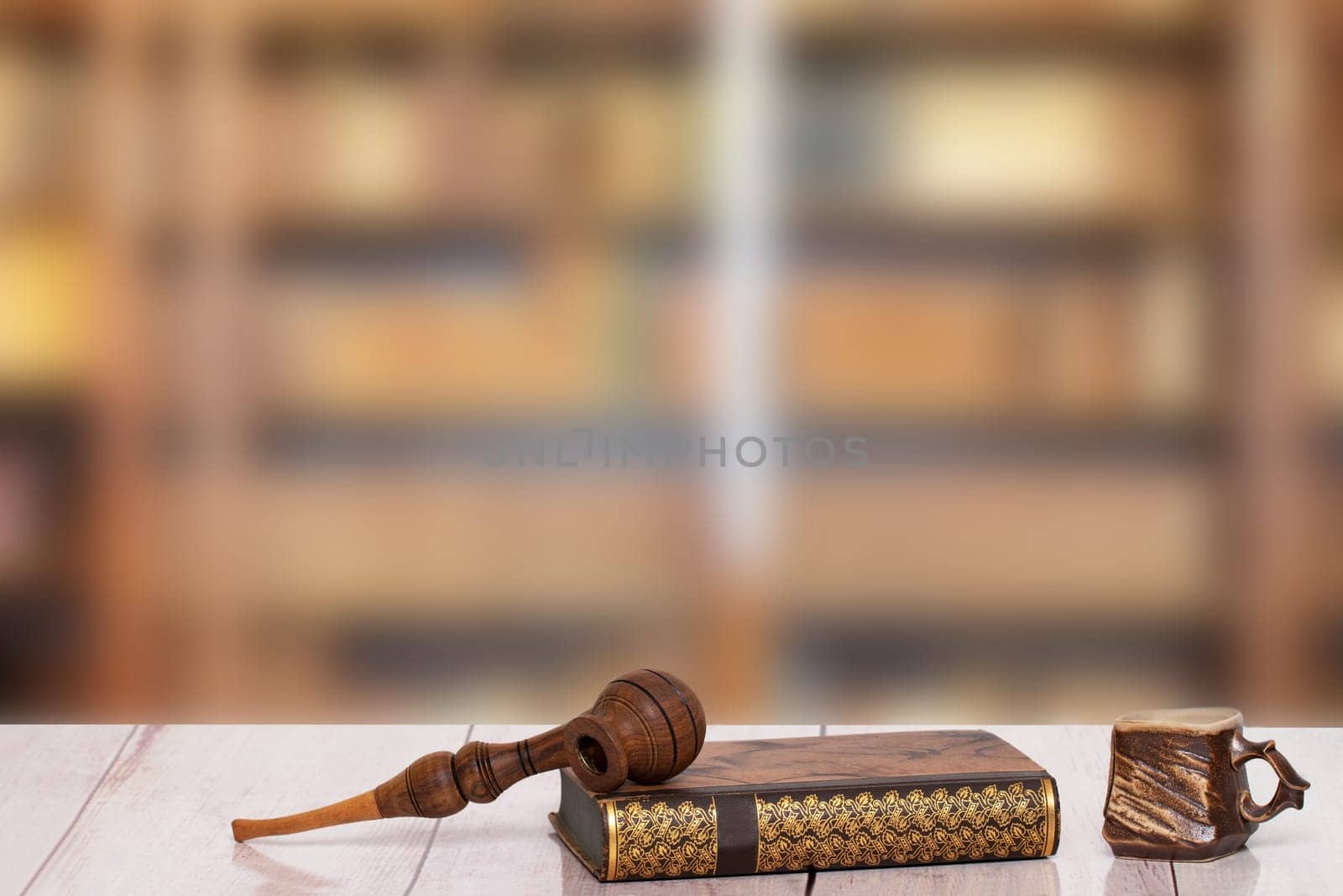 Books backgrounds. Closeup of an antique book with a tobacco pipe on it and a brown clay mug on a light pink desk in front of a blurred bookshelf in a library. Teachers day, back to school. Science and education concept. Space.