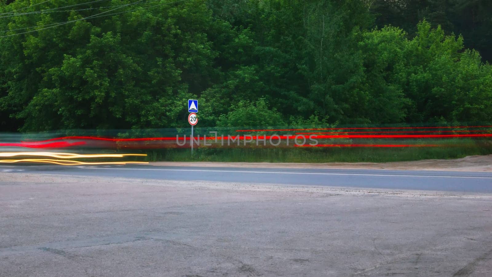 View of the countryside asphalt road with motion blurred glowing traces of car headlights. 20 km speed limit and road hump traffic sign against green trees on the roadside. Selective focus, long exposure.