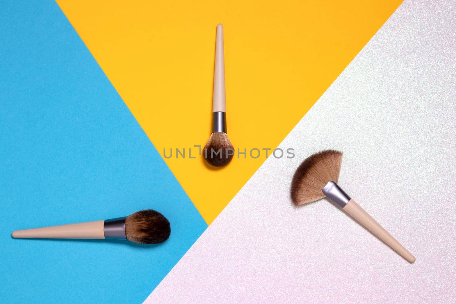 Three professional makeup brushes flat lay on a bright pink, yellow and blue background with copy space. Template for cosmetics product display montage or advertising text. Beauty and skincare concept. Top view.