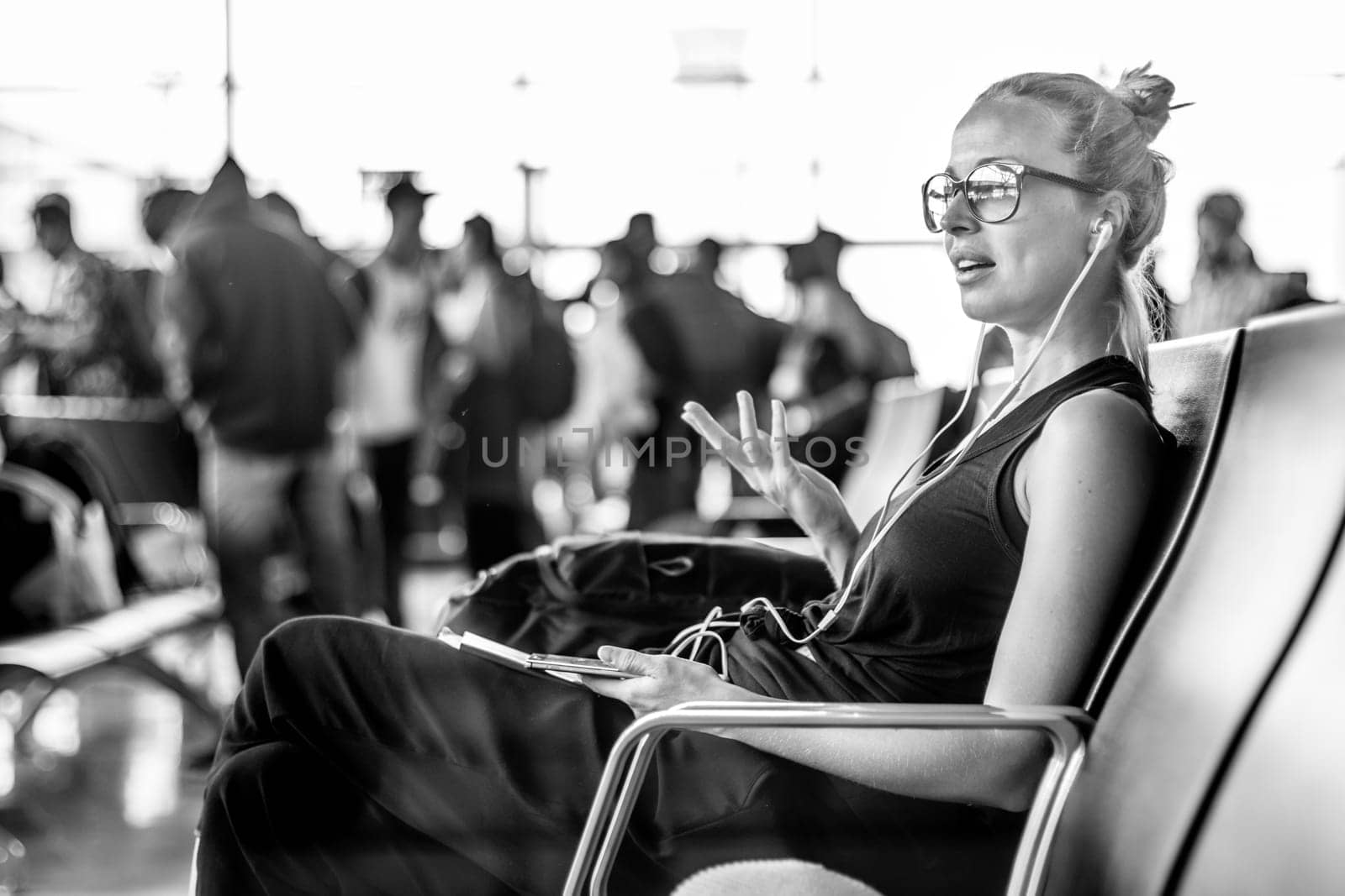 Casual sporty young blond female traveler talking on her cell phone while waiting to board a plane at the departure gates at the asian airport terminal.