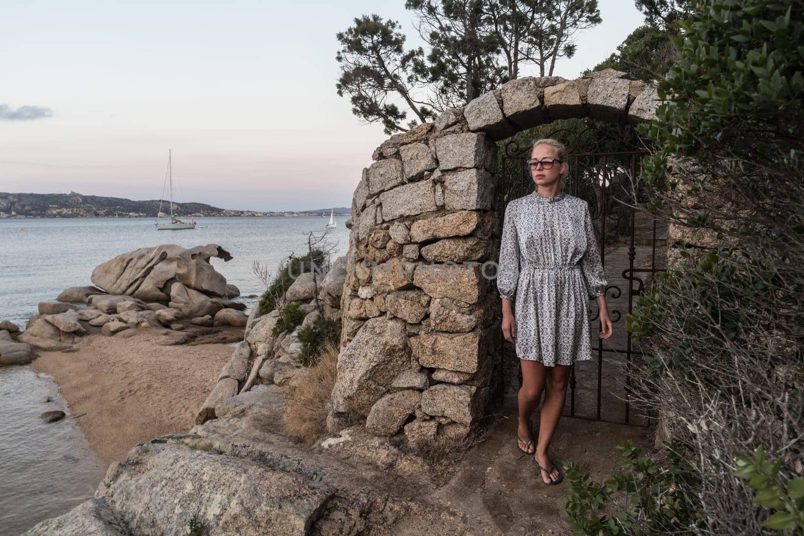 Beautiful woman in luxury summer dress enjoying peaceful seascape of Porto Rafael bay at Mediterranean sea of Costa Smeralda, Sardinia, Italy at dusk.