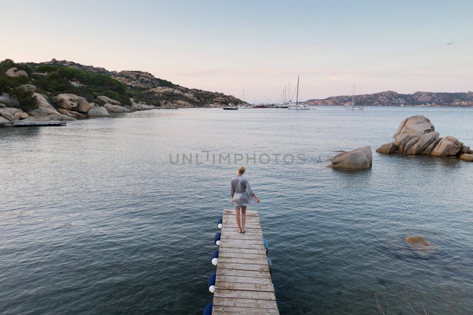 Beautiful woman in luxury summer dress standing on wooden pier enjoying peaceful seascape at dusk. Female traveler stands on a wooden pier in Porto Rafael, Costa Smeralda, Sardinia, Italy.