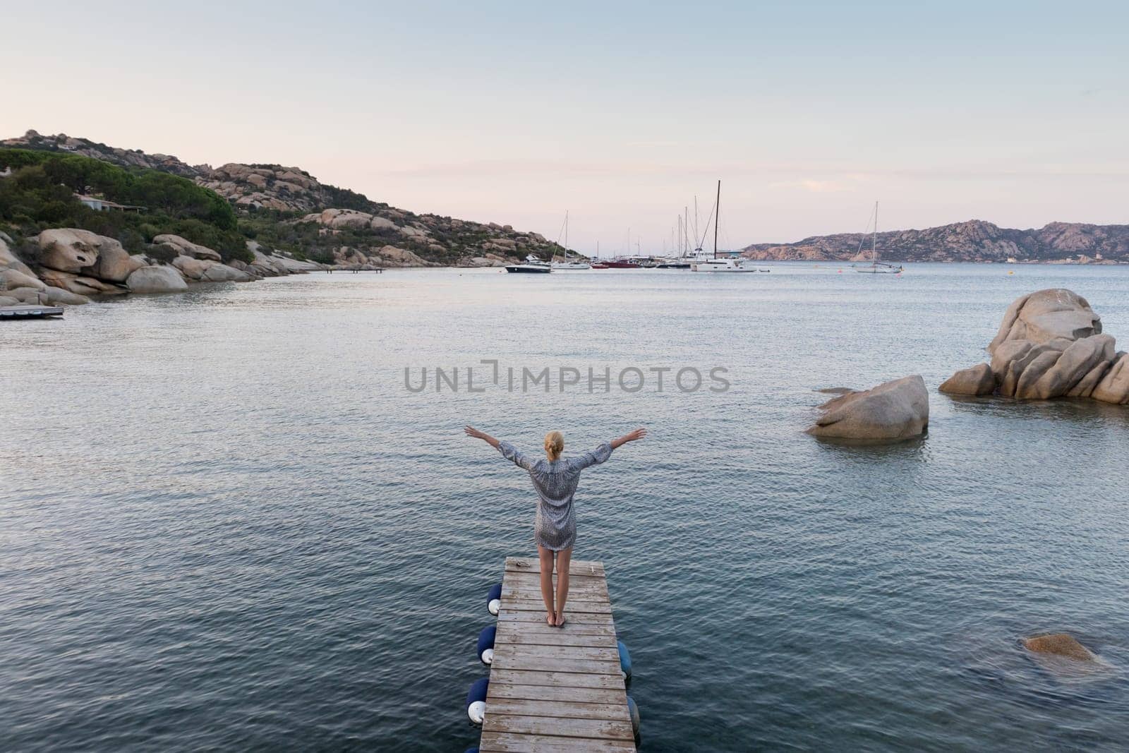 Beautiful woman standing on wooden pier arms rised enjoying peaceful seascape at dusk. Female traveler stands on a wooden pier in Porto Rafael, Costa Smeralda, Sardinia, Italy.