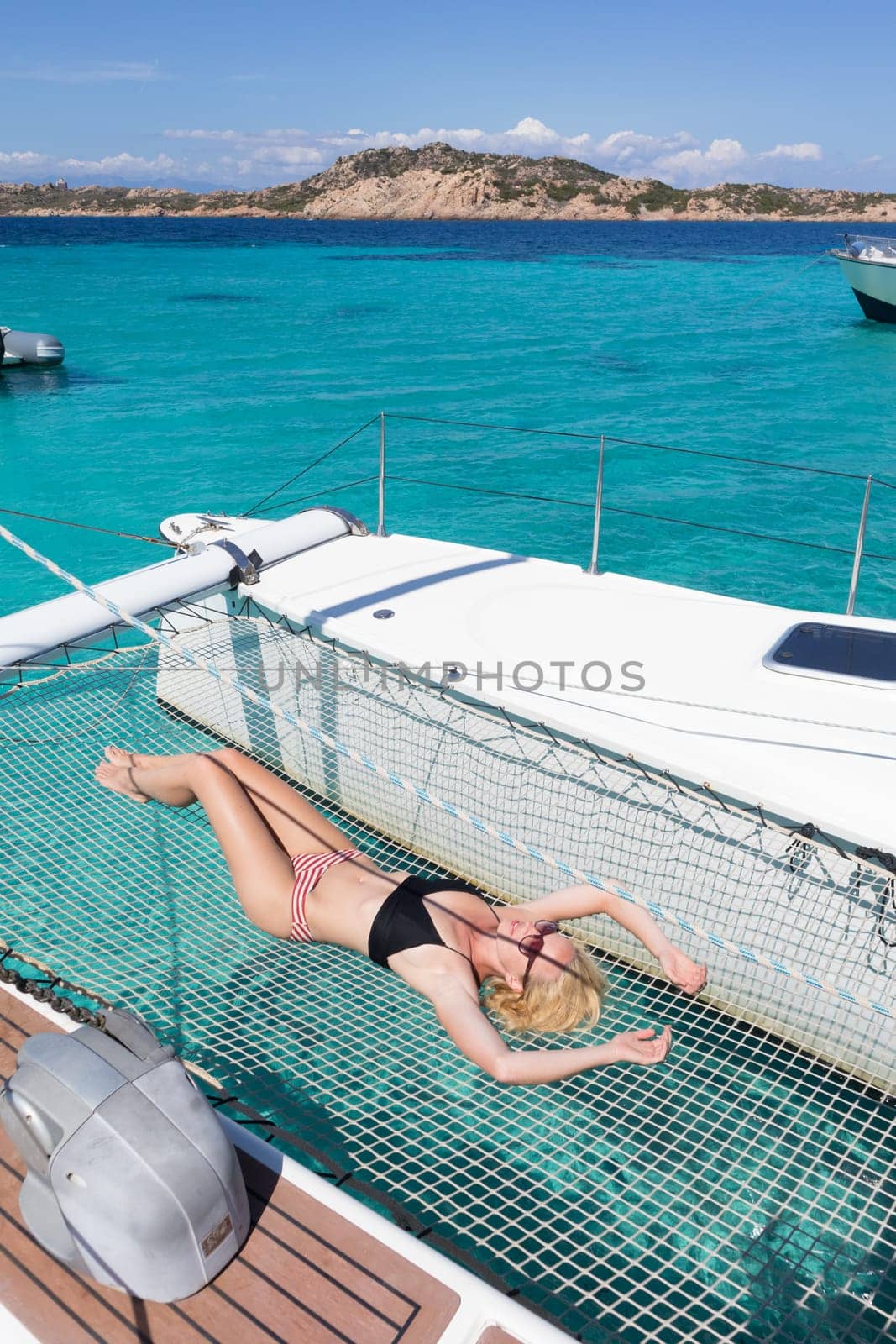 Womanin bikini tanning and relaxing on a summer sailin cruise, lying in hammock of luxury catamaran near picture perfect white sandy beach on Spargi island in Maddalena Archipelago, Sardinia, Italy.