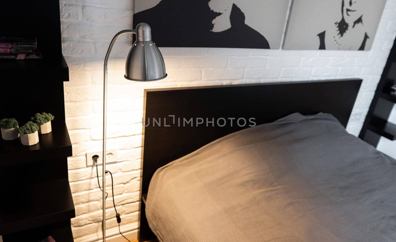Steel downbridge floor lamp with a long arm and opaque shade near bed in black and white modern bedroom. Metal Task Lamp with an adjustable reading light. Stylish bedside lamp in scandinavian interior