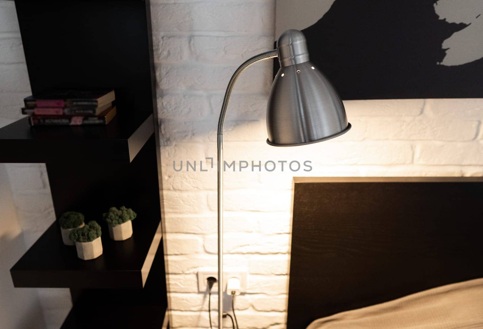 Steel downbridge floor lamp with a long arm and opaque shade near bed in black and white modern bedroom. Metal Task Lamp with an adjustable reading light. Stylish bedside lamp in scandinavian interior