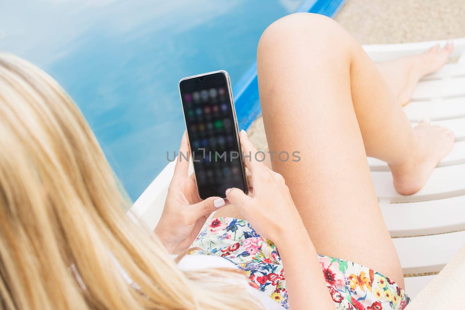 The blond hair woman is using mobile phone at poolside outdoors and using applications.