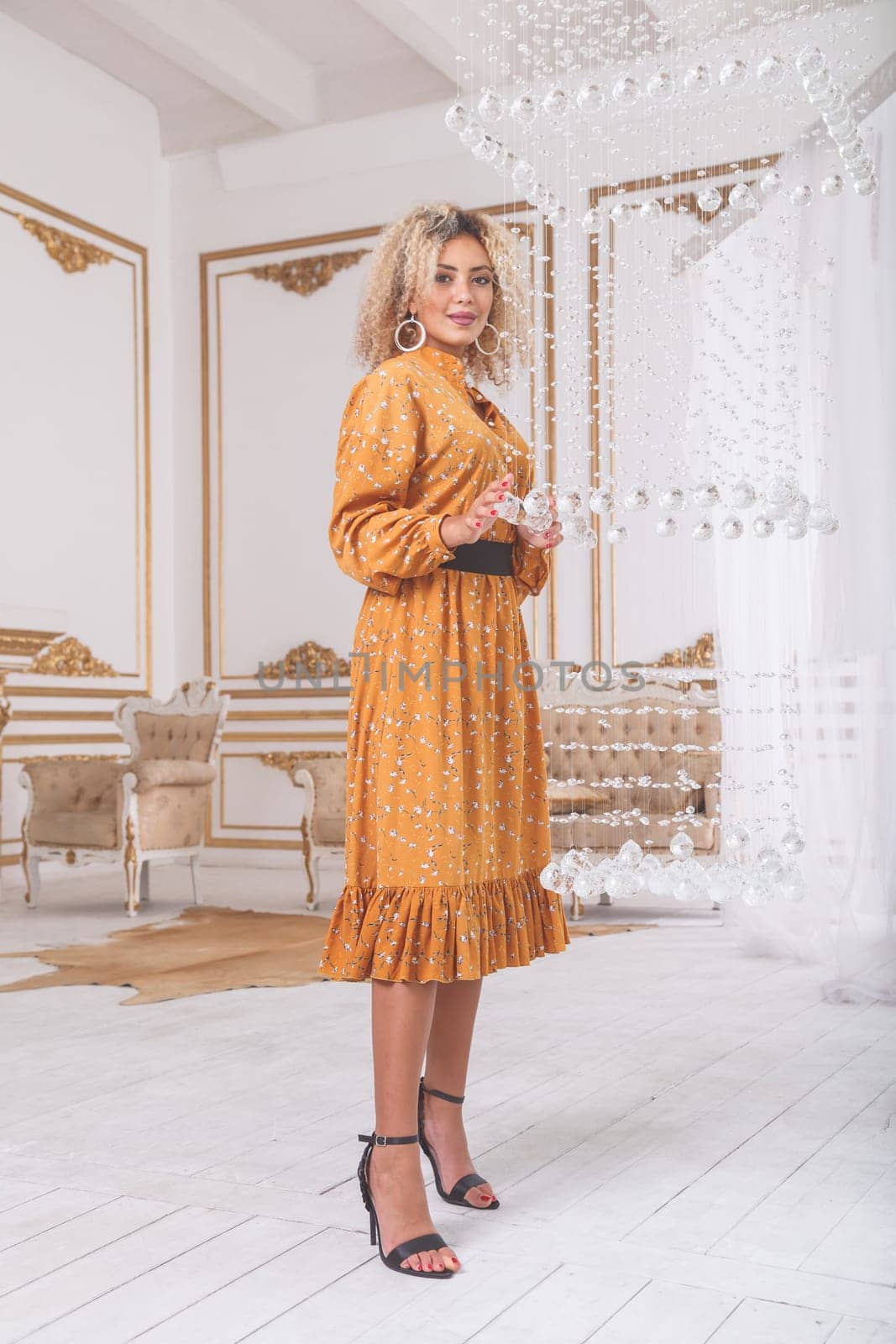 Beautiful african mixed young woman in fashionable dress and heels posing near crystal chandelier in luxury interior