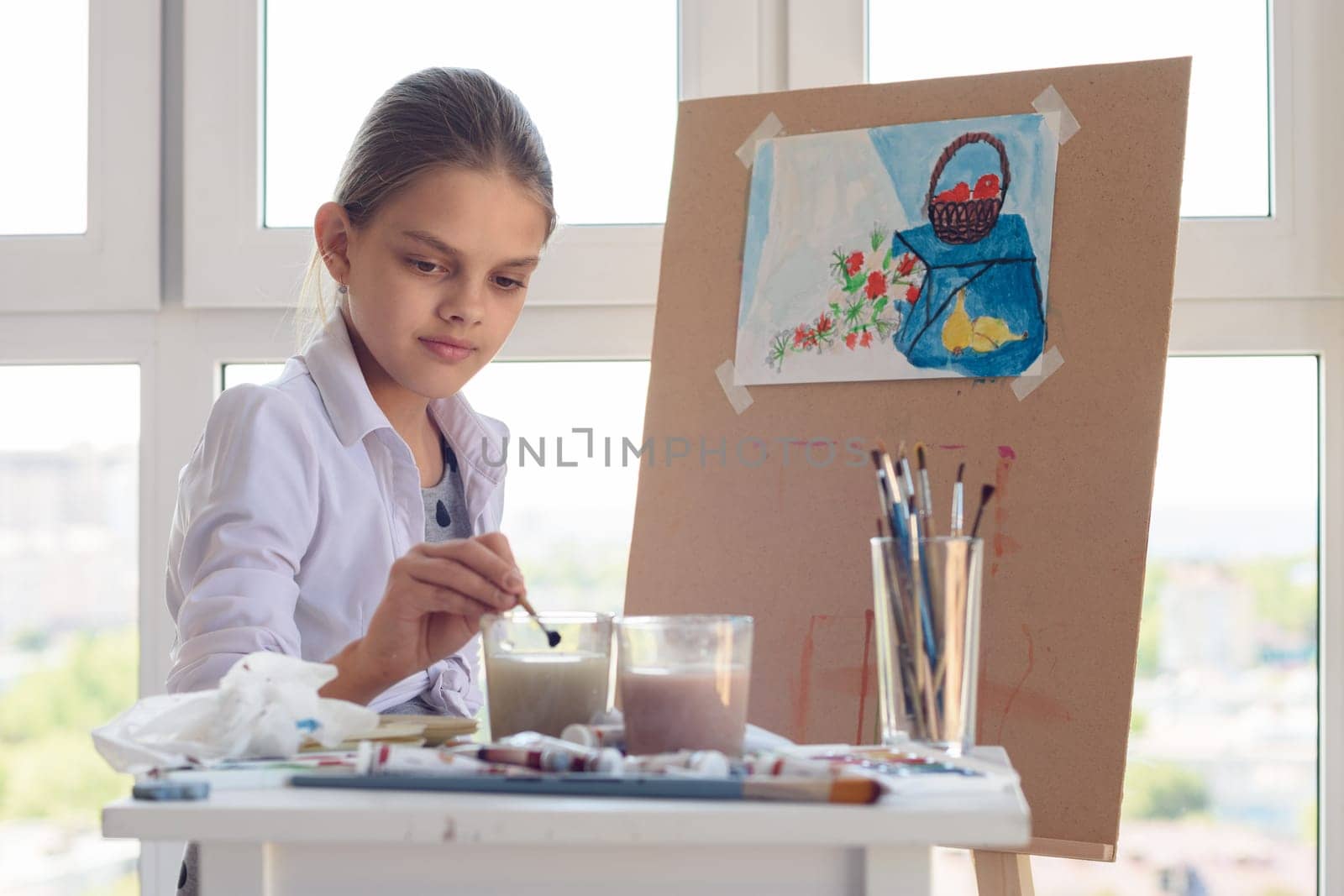 girl sits behind an easel and washes her brush in a glass of water