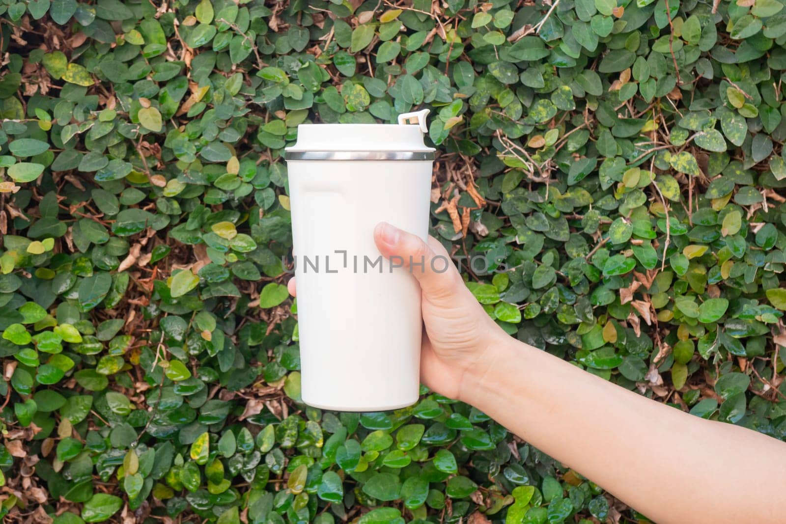Aluminum thermos mug on hand with green leaves wall, stock photo