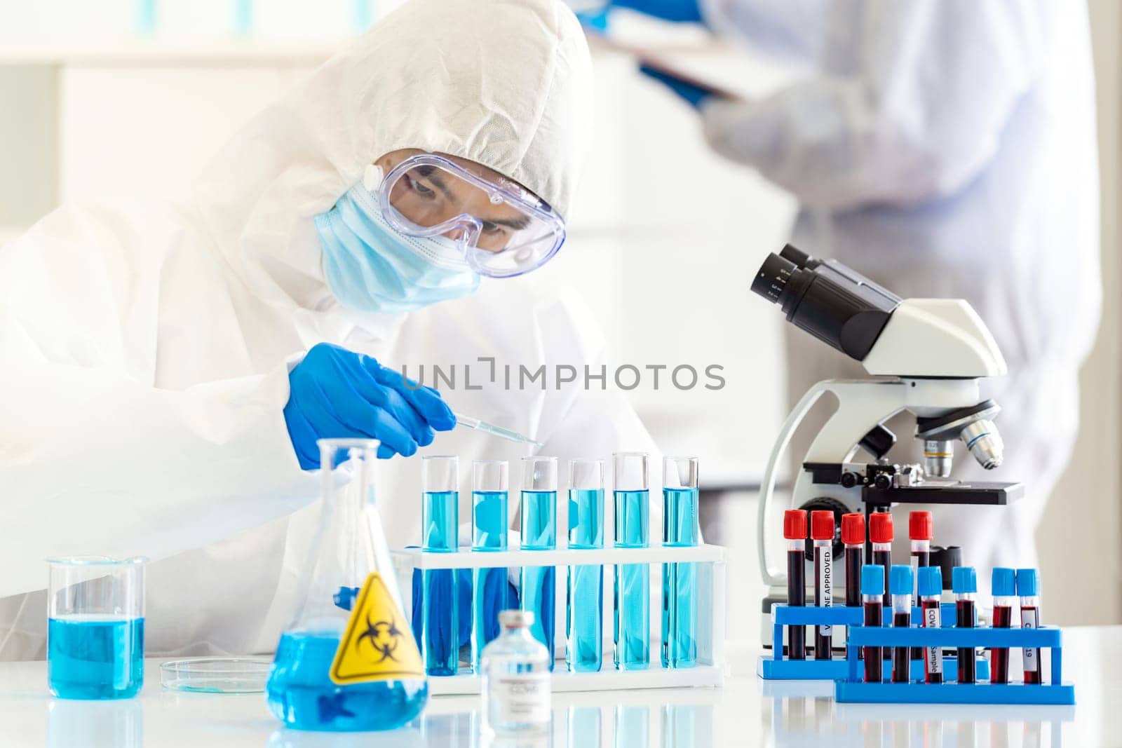 Scientists use dropper to drop chemical reagent to test tube in tube rack to research and develop vaccine for coronavirus covid-19 pandemic with his colleague in background. Medical Science concept.