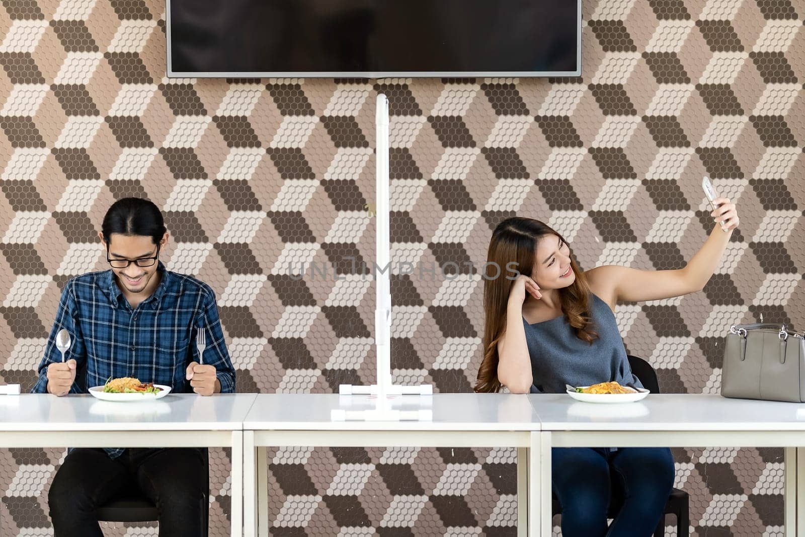 Asian couple take a selfie photo together in new normal social distance restaurant with table shiled partition to reduce infection of coronavirus covid-19 pandemic. Restaurant new normal lifestyle.