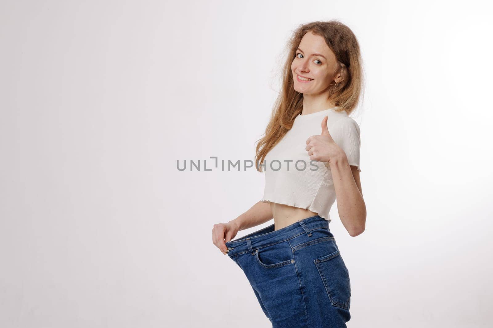Happy woman showing weight loss success by wearing oversized jeans, smiling, and giving a thumbs-up on a white background.