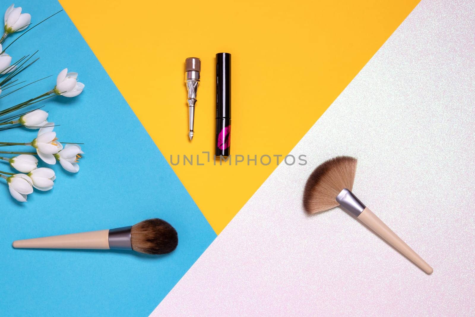 Professional makeup brushes, lipsticks and spring flowers flat lay on a bright pink, yellow and blue background with copy space. Template for cosmetics product display montage or advertising text. Beauty and skincare concept. Top view.