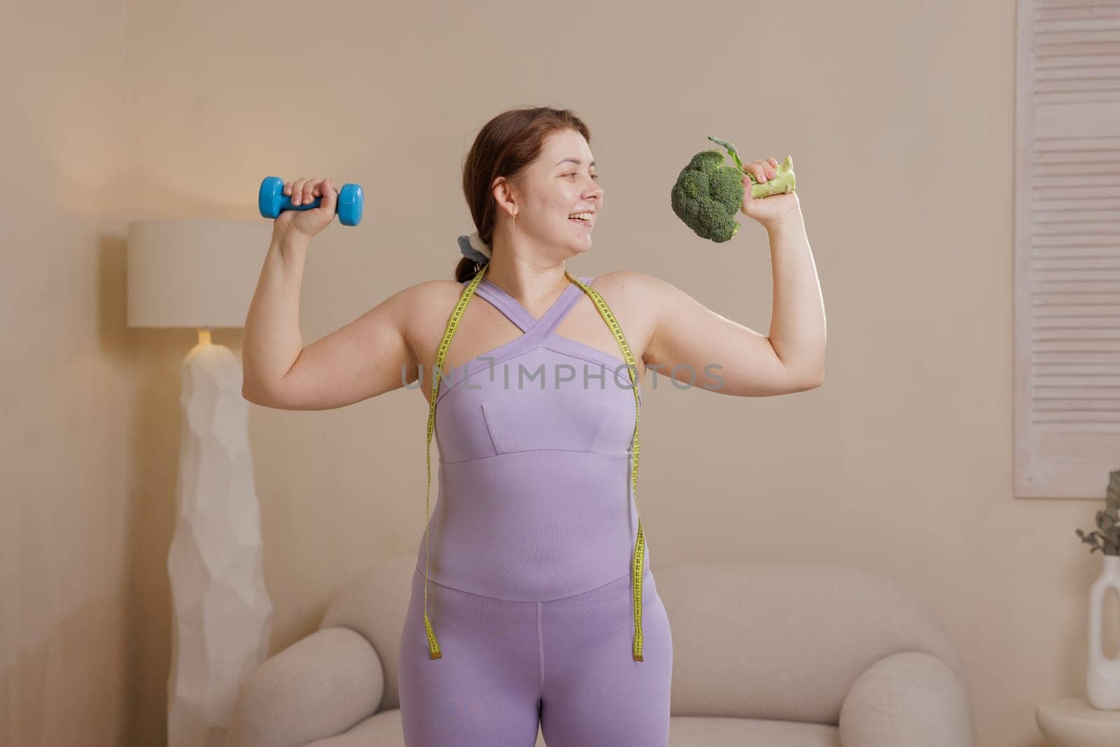 Happy overweight woman in sportswear combining fitness and healthy eating holding dumbbell and broccoli symbolizing well-being, weight loss and body positivity.