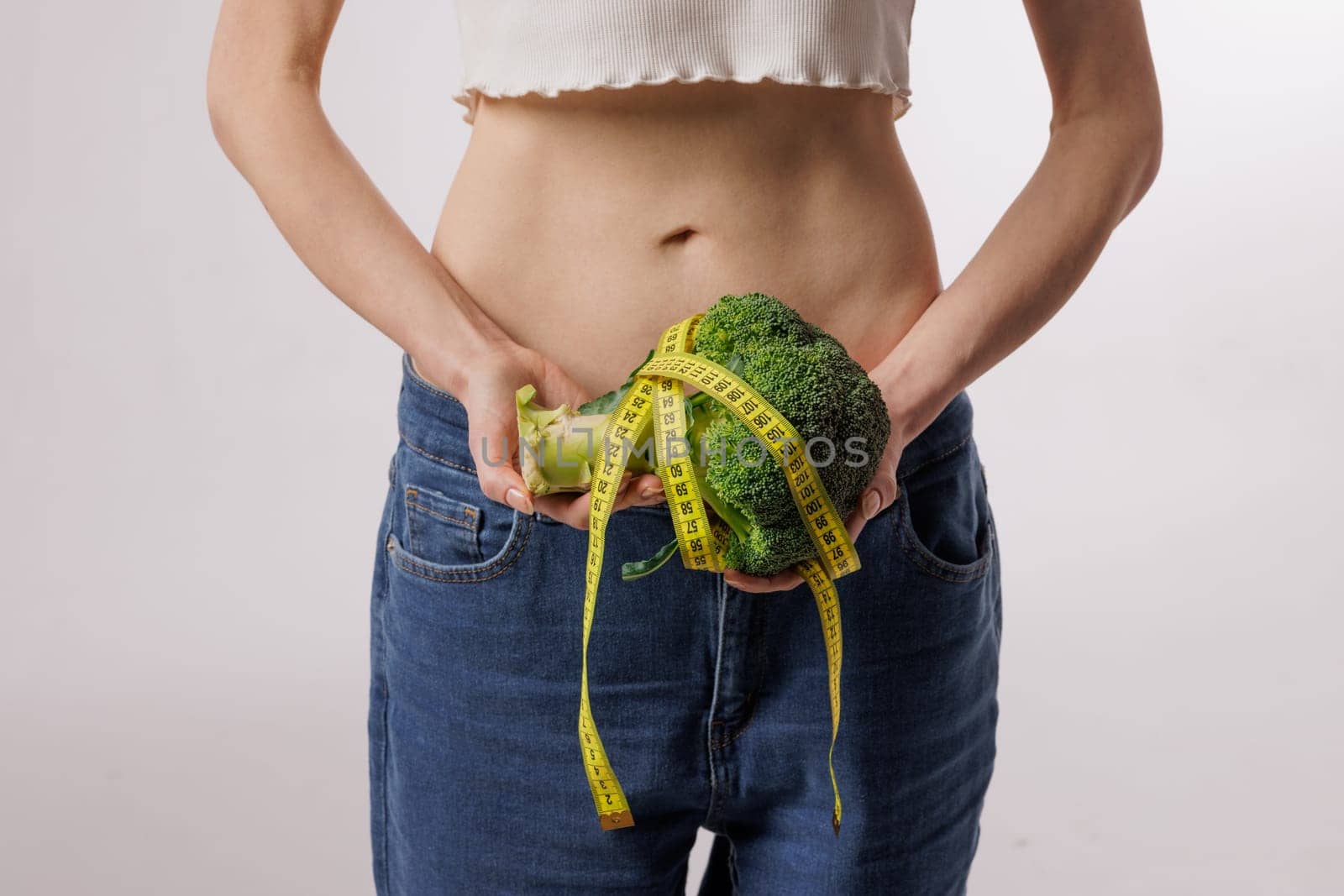 Slim woman holding broccoli wrapped in a measuring tape, symbolizing healthy eating, weight loss, diet, and nutrition on a white background.