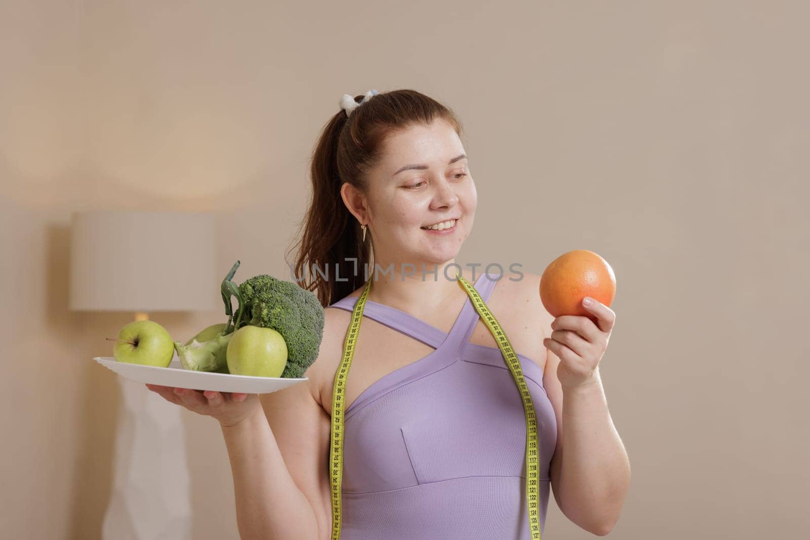 Smiling overweight woman in tracksuit holding healthy fruits and vegetables promoting balanced diet, weight loss and healthy lifestyle.
