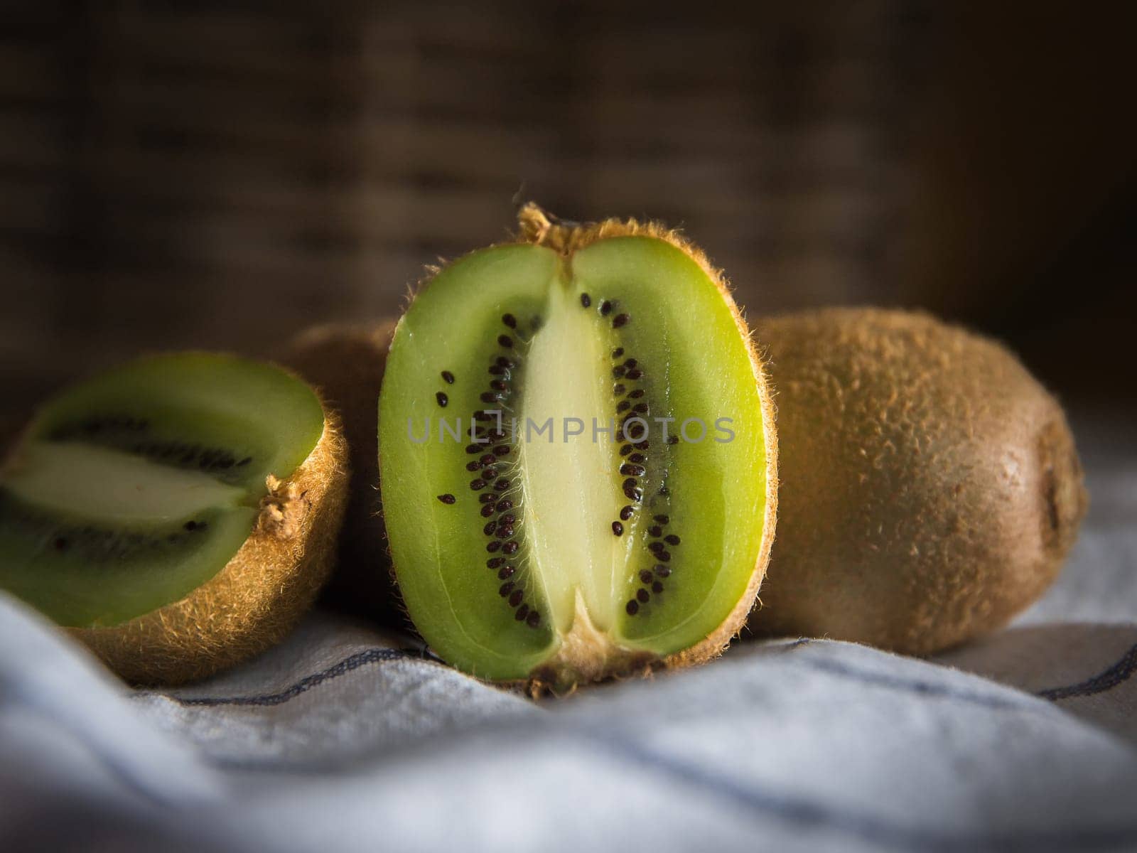 Close-up of a group of kiwis, one of them cut in half. Dark background with a wicker basket. On a white tablecloth with blue lines.