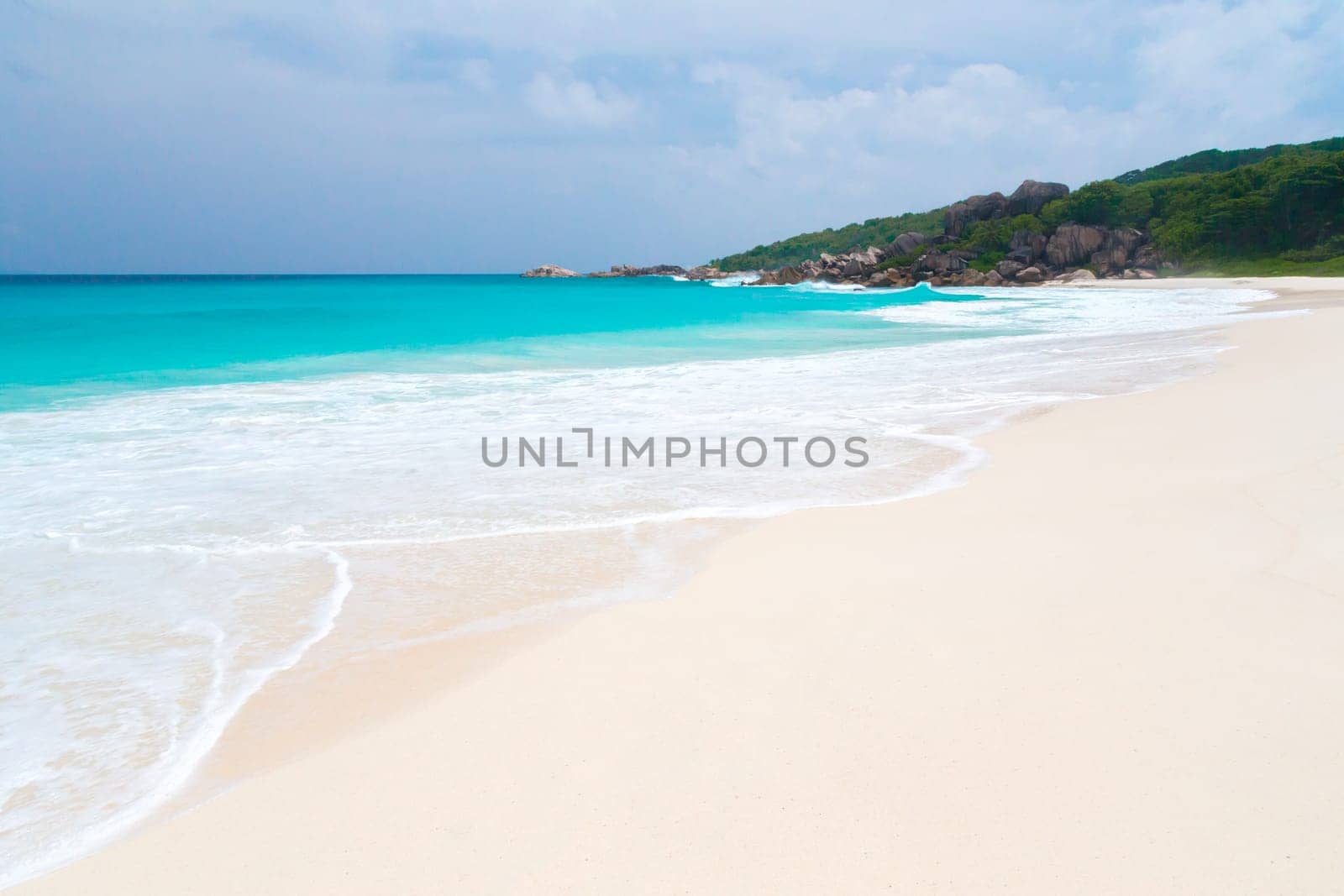 White sand beach with some waves and blue sky in the far a hill with green plants
