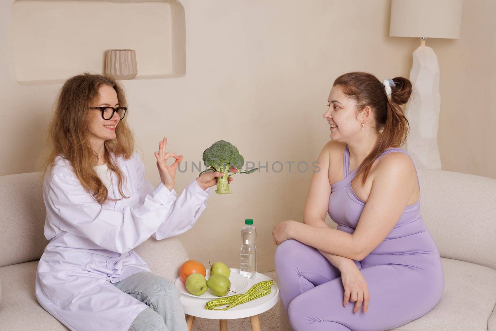 Nutritionist and young woman discussing healthy eating, with fresh fruits, broccoli, and a measuring tape, promoting weight loss, diet, and wellness in a cozy home setting.