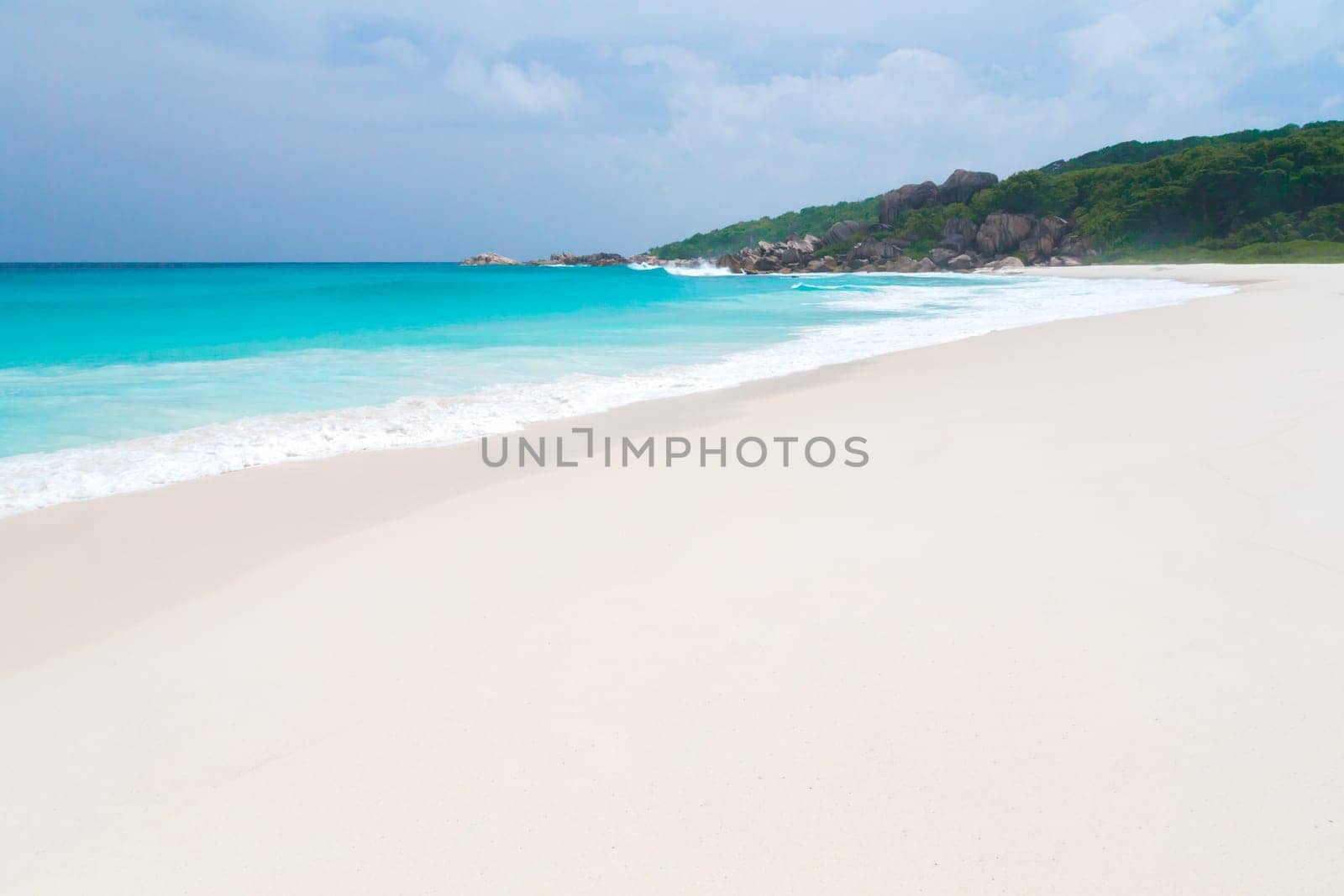 White sand beach with some waves and blue sky in the far a hill with green plants