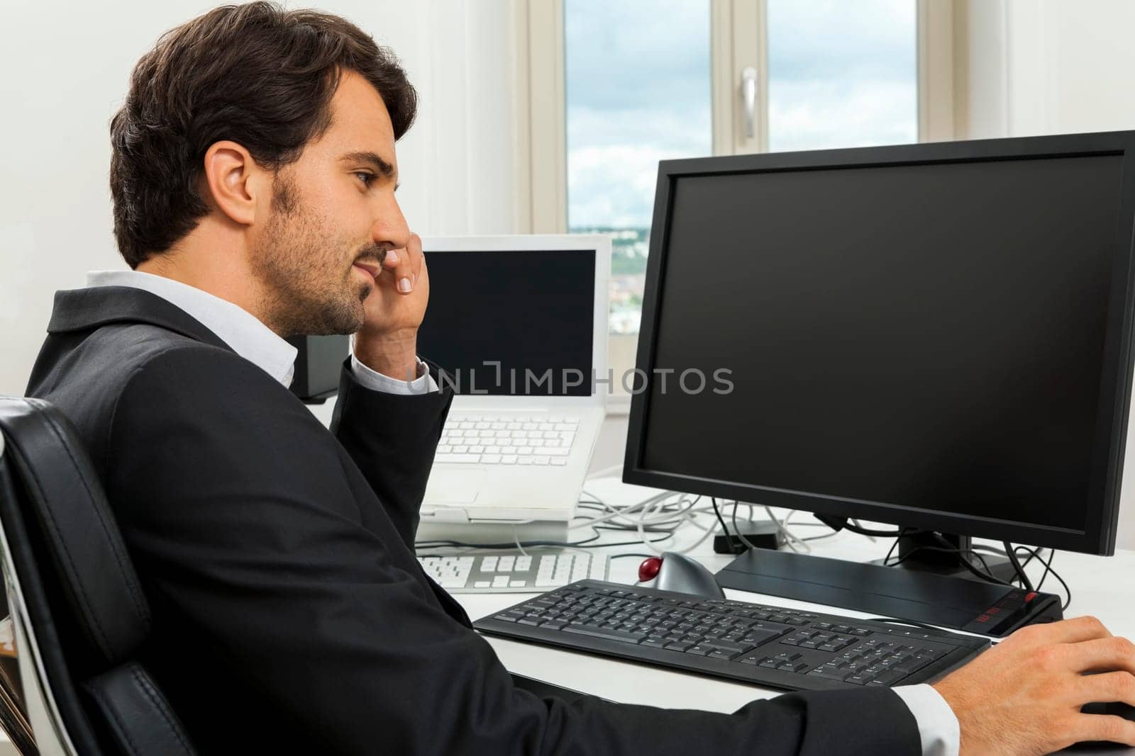 Young successful businessman with black suit in the office at work with computer in concept