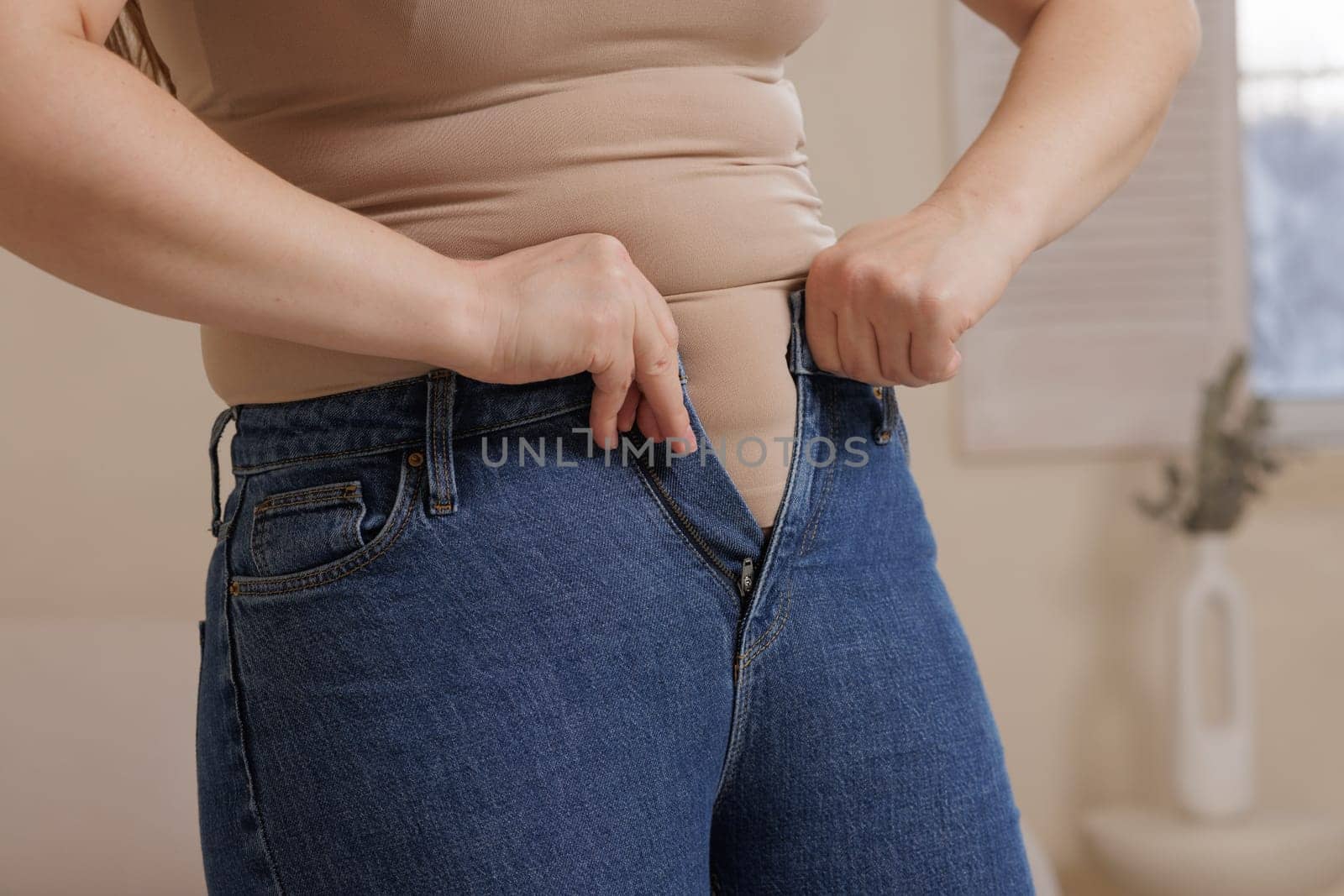 A woman struggles to button tight jeans, highlighting weight gain, body changes, fashion challenges, and body positivity in a relatable moment.