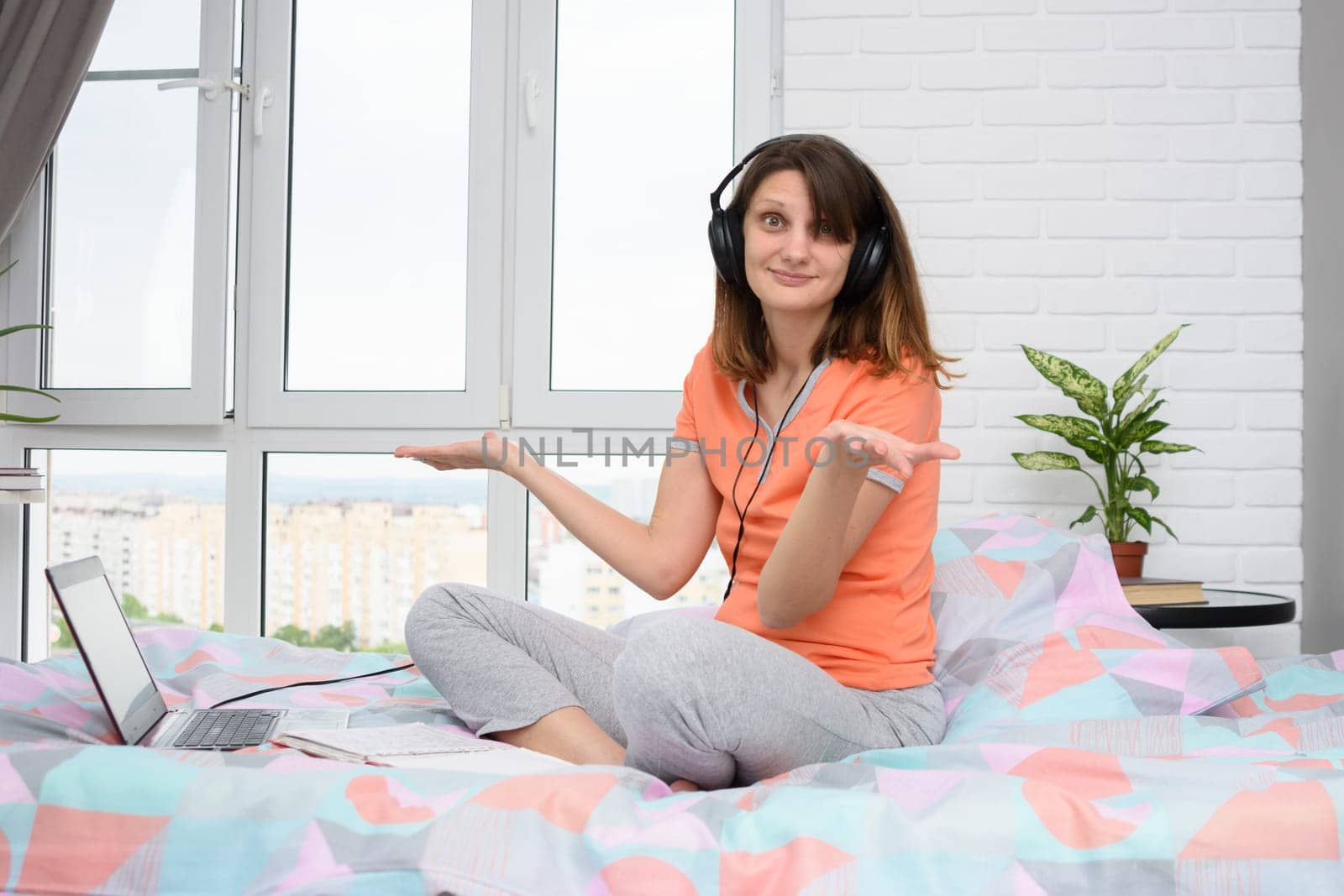 The girl spread her hands in surprise, sitting in headphones and with a laptop on the bed at home