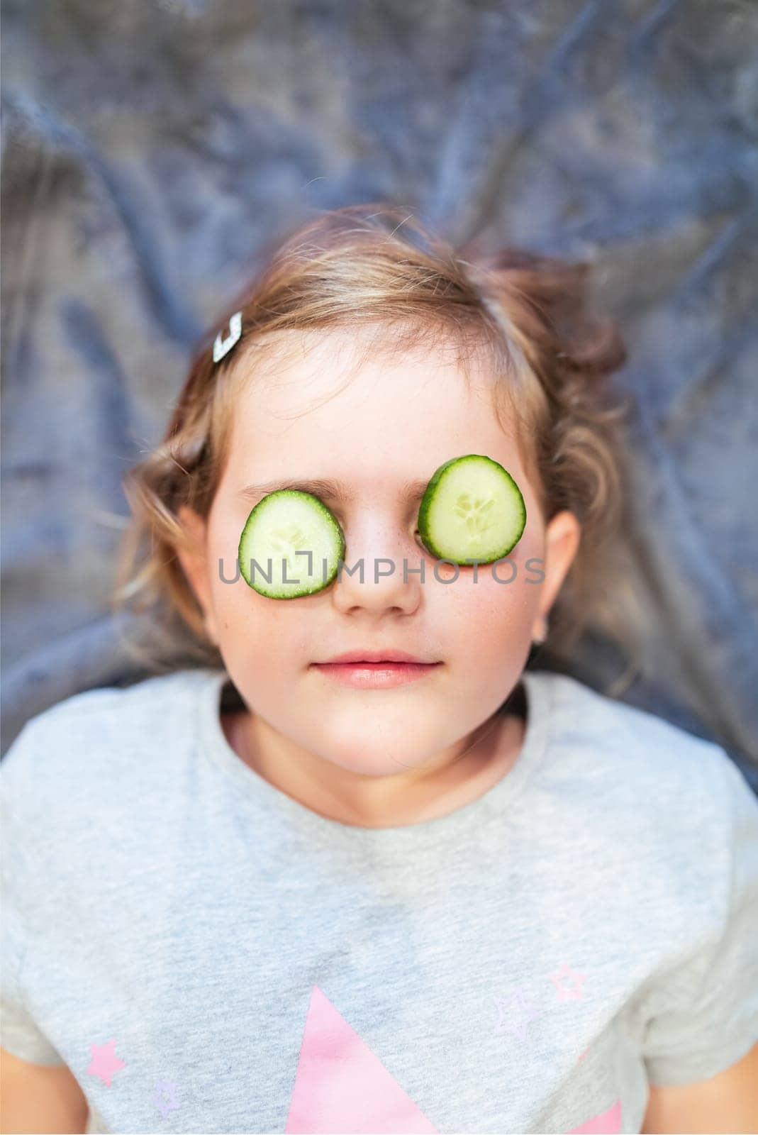 Funny small girl with piece of cucumber on their eyes like a mask, beauty and health concept, indoor closeup portrait