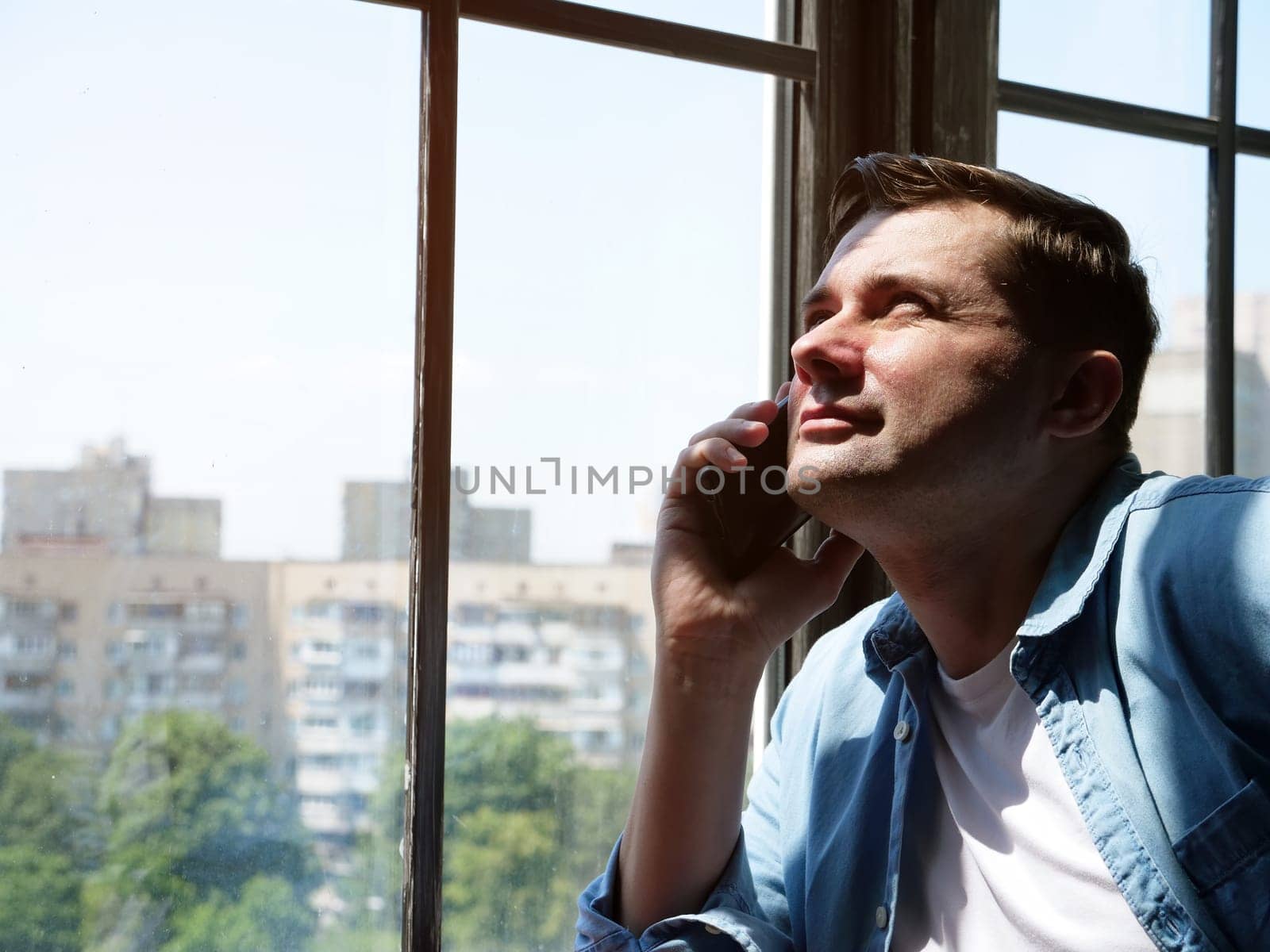 Portrait of a young man who stands by the window and makes a phone call. Communication concept.