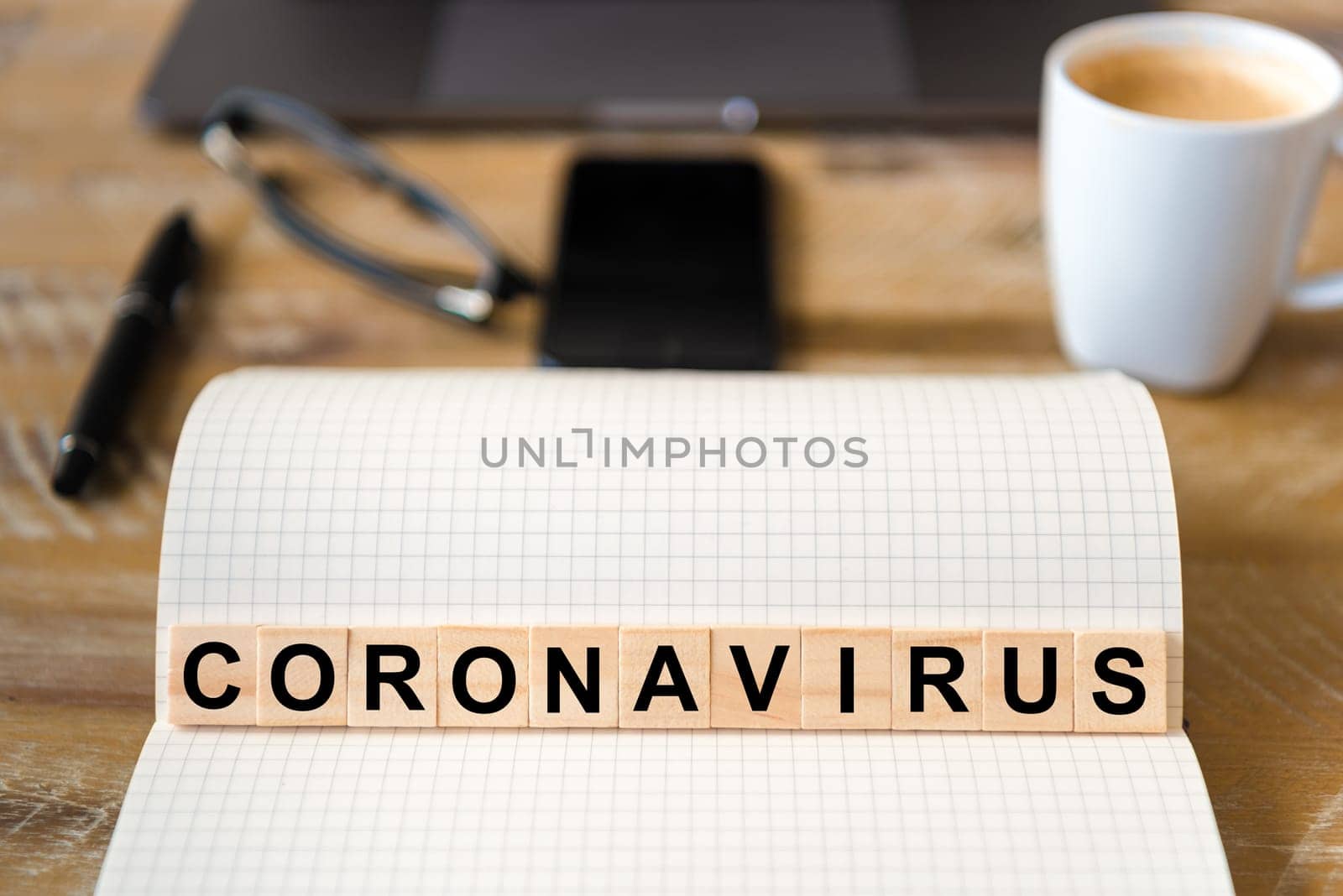 Closeup on notebook over wood table background, focus on wooden blocks with letters making Coronavirus writing. Business concept with laptop, glasses, pen and mobile phone in a defocused background