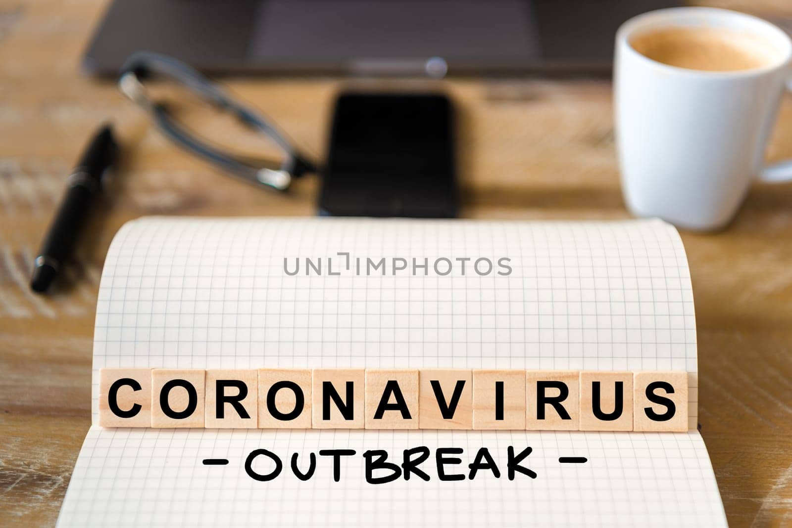 Closeup on notebook over wood table background, focus on wooden blocks with letters making Coronavirus Outbreak writing. Business concept with laptop and mobile phone in a defocused background