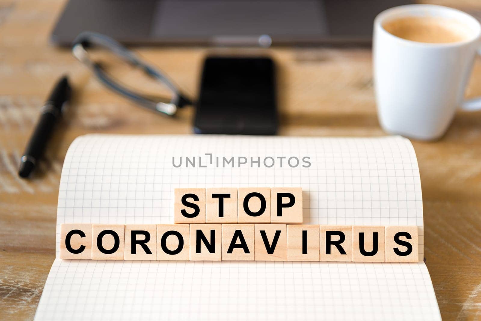 Closeup on notebook over wood table background, focus on wooden blocks with letters making Stop Coronavirus writing. Business concept with laptop and mobile phone in a defocused background